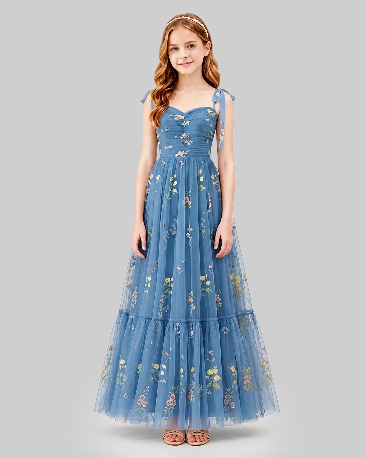 A girl wearing the Dideyttawl Embroidered Tulle Formal Dress for Girls with Tie Straps stands facing forward. The long blue couture gown features delicate floral embroidery. She has long, wavy hair adorned with a headband and is wearing sandals. The background is plain gray. Main image